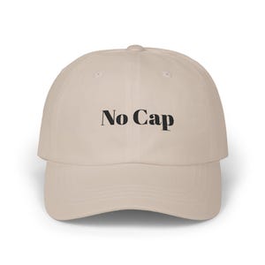 May include: A beige baseball cap featuring the text No Cap embroidered in black thread on the front panel. The hat has a structured crown, a curved brim, and a button at the top center. The design is centered and clearly visible on the fabric.