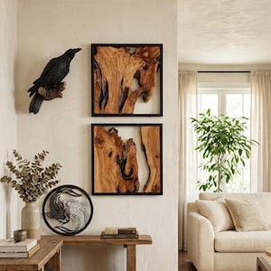 May include: A black raven sculpture perched on a branch, mounted on a white wall. Below, two framed wood art pieces with black borders hang. A wooden table displays a decorative plate with a yin-yang koi fish design and a vase with dried foliage.