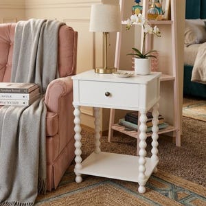 May include: A white side table with a drawer and a lower shelf. The table has decorative, turned legs and a dark knob. A gold lamp with a beige shade sits on top. The table is in a room with a pink chair and a patterned rug.