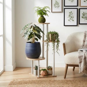 May include: A tiered plant stand featuring white metal supports and light wood circular shelves. The stand holds a green kokedama, a large alocasia in a blue ceramic pot, a trailing succulent in a gold pot, and three small potted plants at the base.