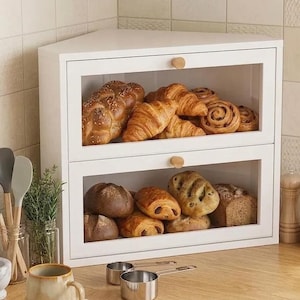 May include: A white corner bread box with two tiers, featuring clear glass front panels and small wooden knobs. The unit is filled with various baked goods, including braided bread, croissants, cinnamon rolls, and artisan loaves of bread.