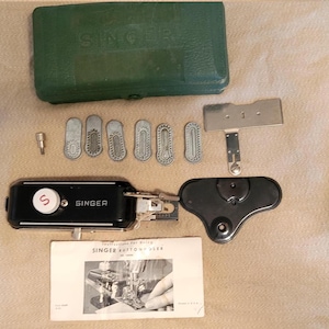 May include: A vintage Singer buttonholer attachment set including a green plastic storage case, a black metal buttonholer unit with a white dial, six metal cams, a metal throat plate, a black cover piece, and an instruction manual.