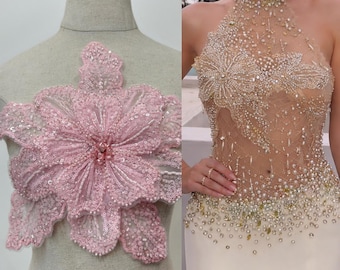 May include: A large, light pink floral applique featuring intricate beadwork and sequins on a mesh base, displayed on a mannequin, alongside a sheer, high-neck bodice embellished with gold beads, pearls, and sequins on a light fabric base.