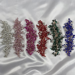 May include: Six jewelled hair accessories in silver, pink, magenta, red, green, and blue. Each piece has a floral vine design with sparkling crystals. They are displayed on a white satin background.