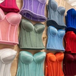 May include: A collection of colorful corsets in various shades, including pink, purple, blue, red, and green. The corsets have a structured design with boning and a sweetheart neckline. Some corsets have a sheer overlay, and others are made of satin.