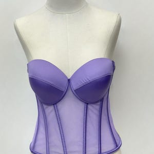 May include: A lavender strapless corset displayed on a mannequin. The bodice features a structured design with a sheer panel and vertical boning. The bust cups are a darker shade of lavender, creating a contrast. The corset is a fashionable garment.