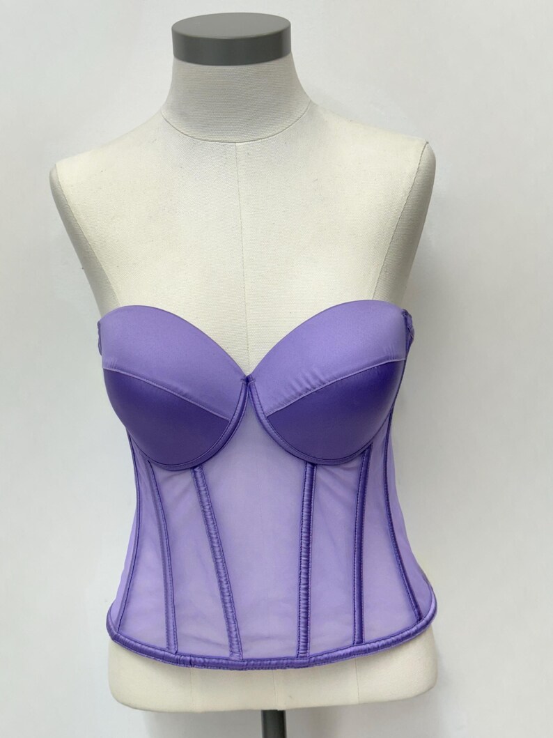 May include: A lavender strapless corset displayed on a mannequin. The bodice features a structured design with a sheer panel and vertical boning. The bust cups are a darker shade of lavender, creating a contrast. The corset is a fashionable garment.