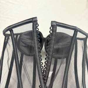 May include: Black corset with a sheer mesh overlay. The corset features a structured design with vertical boning and a decorative trim along the edges. The cups are made of a solid black material.
