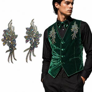 Puede incluir: Un chaleco de terciopelo verde oscuro con adornos florales y una pajarita a juego. Dos broches florales con piedras iridiscentes se muestran sobre un fondo blanco. El chaleco se combina con una camisa y un pantalón negros.