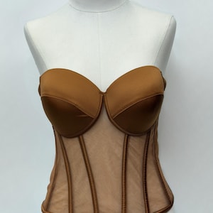 May include: A strapless brown corset with a sweetheart neckline. The bodice is made of a sheer, nude-colored mesh with vertical brown boning details. The bust is a solid, satiny brown material.