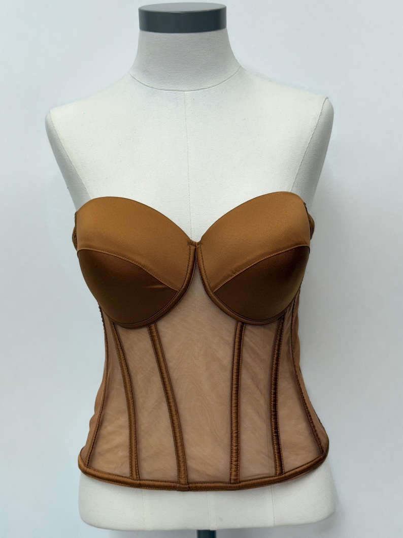 May include: A strapless brown corset with a sweetheart neckline. The bodice is made of a sheer, nude-colored mesh with vertical brown boning details. The bust is a solid, satiny brown material.