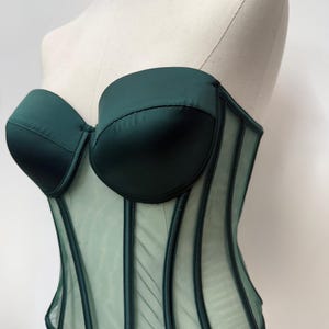 May include: A dark green corset with a sweetheart neckline and boning detail. The bodice is made of a satin-like material, and the lower portion is a sheer mesh. The corset is displayed on a mannequin.