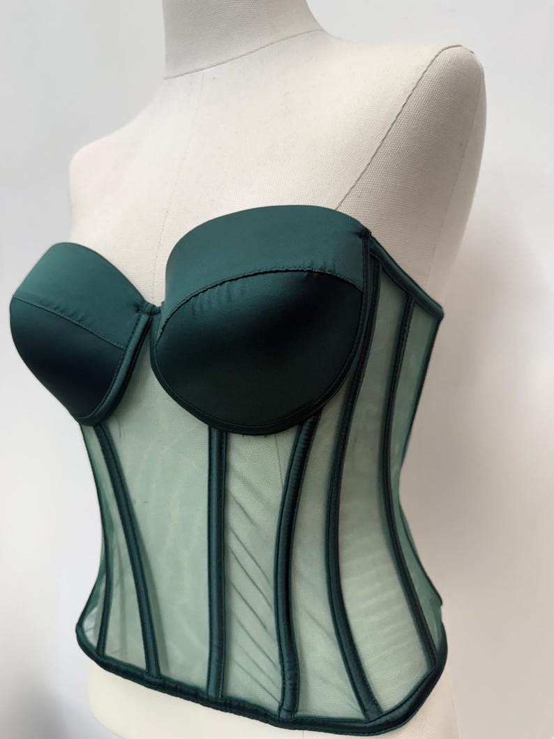 May include: A dark green corset with a sweetheart neckline and boning detail. The bodice is made of a satin-like material, and the lower portion is a sheer mesh. The corset is displayed on a mannequin.