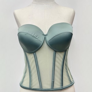 May include: A strapless, sage green corset top displayed on a mannequin. The bodice features a structured design with a sheer mesh panel and vertical boning. The bust is a solid, rounded shape.
