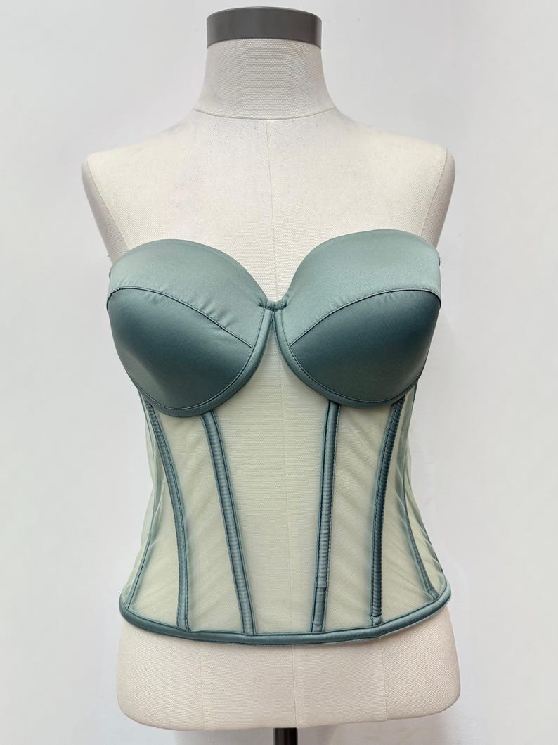 May include: A strapless, sage green corset top displayed on a mannequin. The bodice features a structured design with a sheer mesh panel and vertical boning. The bust is a solid, rounded shape.