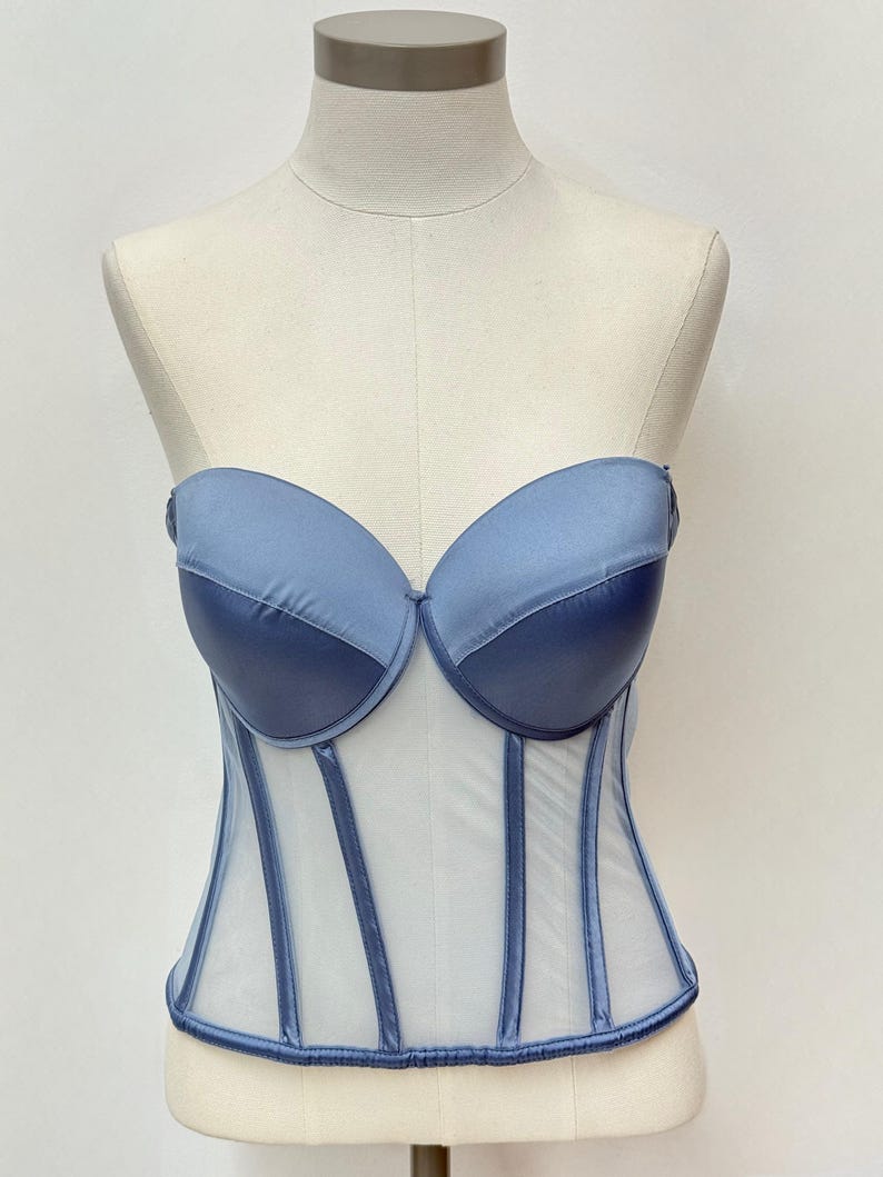 May include: A strapless, blue corset with a sheer, white mesh body. The bust is a solid, darker blue, and the boning is a lighter blue. The corset is displayed on a mannequin.