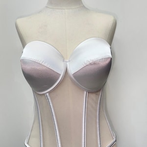 May include: A strapless corset in white and mauve satin, with a sheer mesh lower section. The bust cups are white, transitioning to mauve. Vertical silver-toned boning details the front.