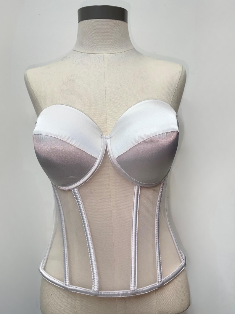 May include: A strapless corset in white and mauve satin, with a sheer mesh lower section. The bust cups are white, transitioning to mauve. Vertical silver-toned boning details the front.