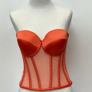 May include: An orange strapless corset with a sweetheart neckline. The bodice features a sheer mesh panel with vertical boning and a satin trim. The corset is displayed on a mannequin.