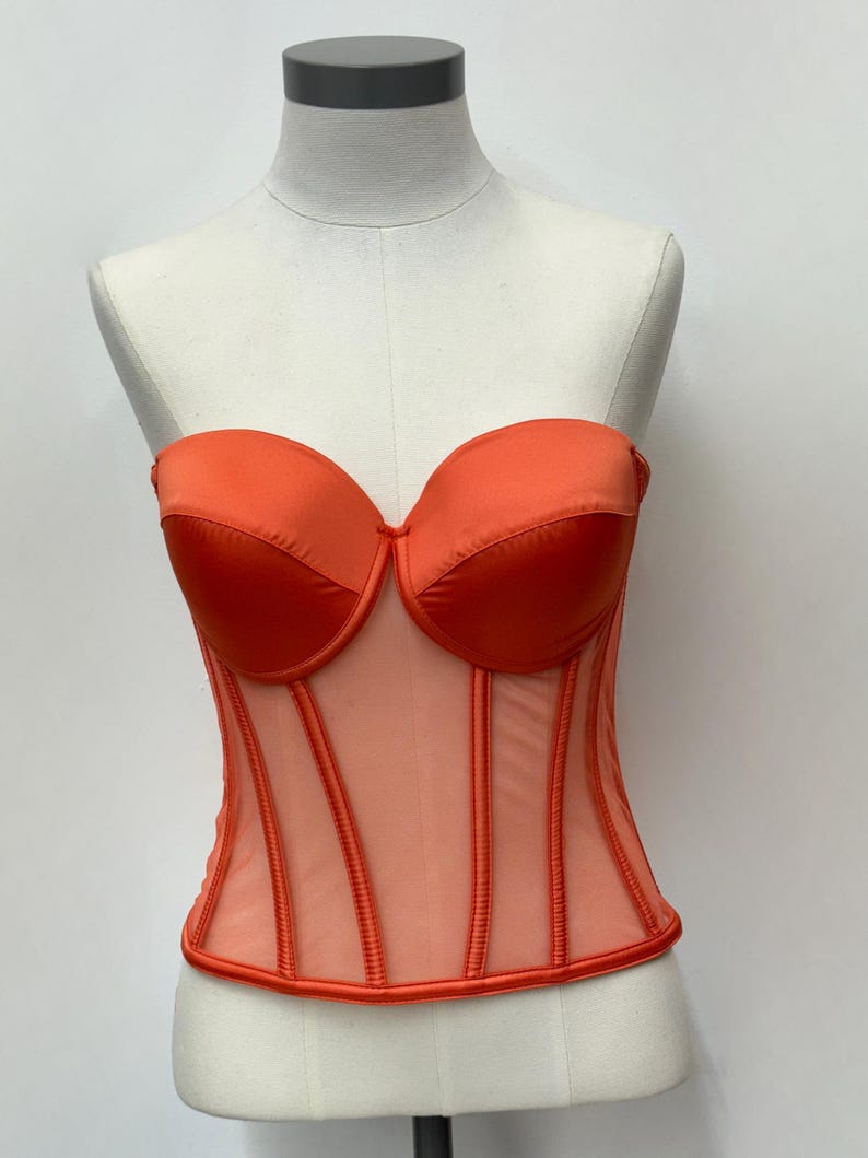 May include: An orange strapless corset with a sweetheart neckline. The bodice features a sheer mesh panel with vertical boning and a satin trim. The corset is displayed on a mannequin.