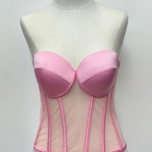 May include: A strapless pink corset displayed on a mannequin. The bust is a darker pink, with a lighter pink mesh body. Vertical seams and trim are a darker pink. The corset is a lingerie item.