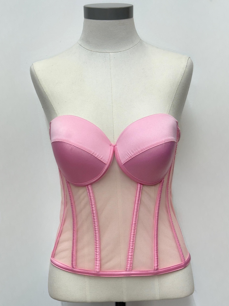 May include: A strapless pink corset displayed on a mannequin. The bust is a darker pink, with a lighter pink mesh body. Vertical seams and trim are a darker pink. The corset is a lingerie item.