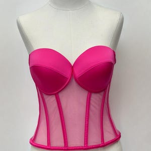 May include: A vibrant fuchsia corset displayed on a mannequin. The strapless top features a sweetheart neckline and structured cups. Vertical boning details the sheer mesh bodice. The corset is a bold, eye-catching piece.