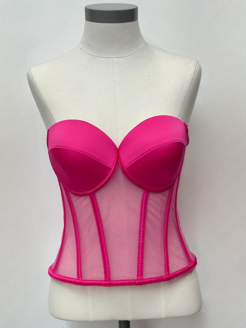 May include: A vibrant fuchsia corset displayed on a mannequin. The strapless top features a sweetheart neckline and structured cups. Vertical boning details the sheer mesh bodice. The corset is a bold, eye-catching piece.