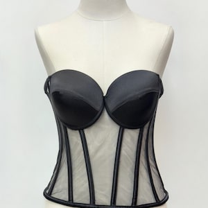 May include: Black bustier corset with a sheer mesh panel and vertical black trim. The top features a sweetheart neckline and is displayed on a mannequin. The corset is a lingerie item.