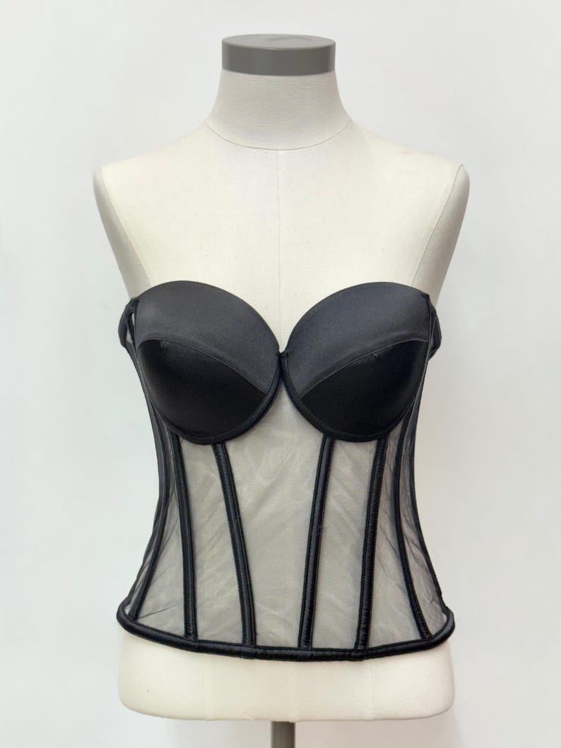 May include: Black bustier corset with a sheer mesh panel and vertical black trim. The top features a sweetheart neckline and is displayed on a mannequin. The corset is a lingerie item.