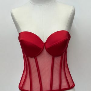 May include: A red corset top with a sweetheart neckline and boning details. The bodice is made of a sheer mesh material with vertical red accents. The top is displayed on a mannequin.