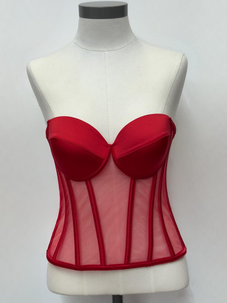 May include: A red corset top with a sweetheart neckline and boning details. The bodice is made of a sheer mesh material with vertical red accents. The top is displayed on a mannequin.