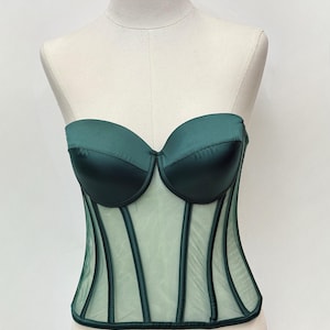 May include: A dark green corset displayed on a mannequin. The bodice features a sweetheart neckline and structured boning. The upper portion is a solid, satin-like material, while the lower section is a sheer mesh with vertical accents.
