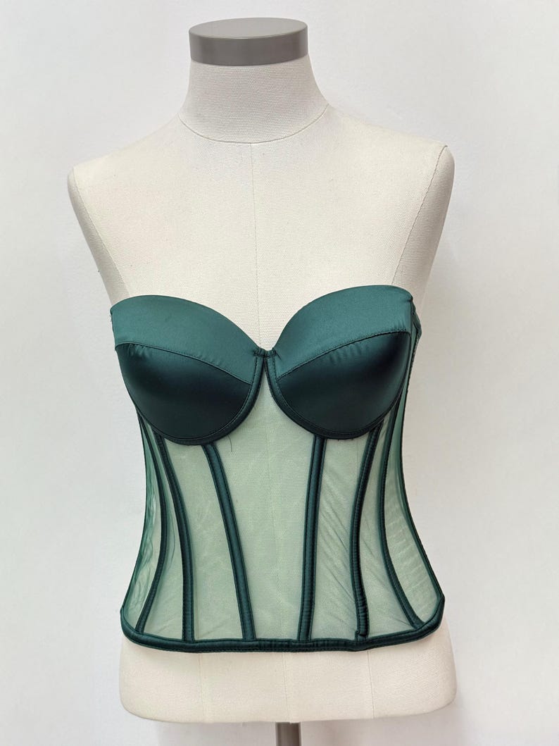 May include: A dark green corset displayed on a mannequin. The bodice features a sweetheart neckline and structured boning. The upper portion is a solid, satin-like material, while the lower section is a sheer mesh with vertical accents.