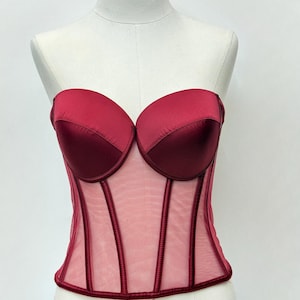 May include: A strapless burgundy corset with a sweetheart neckline. The bodice features a sheer mesh panel with vertical burgundy boning details. The bust is made of a satin-like material. The corset is displayed on a mannequin.