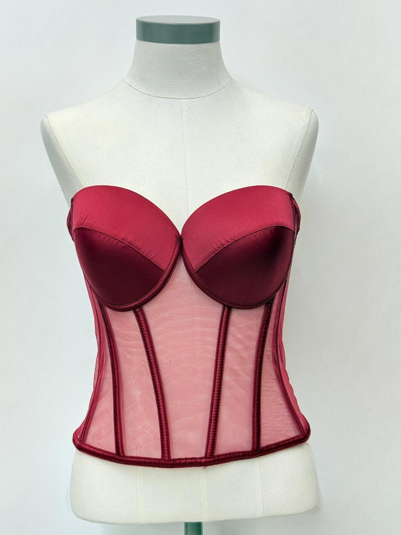May include: A strapless burgundy corset with a sweetheart neckline. The bodice features a sheer mesh panel with vertical burgundy boning details. The bust is made of a satin-like material. The corset is displayed on a mannequin.