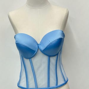 May include: A light blue corset with a sweetheart neckline and boning detail. The bodice is made of a sheer, white fabric with vertical blue accents. The corset is displayed on a mannequin.