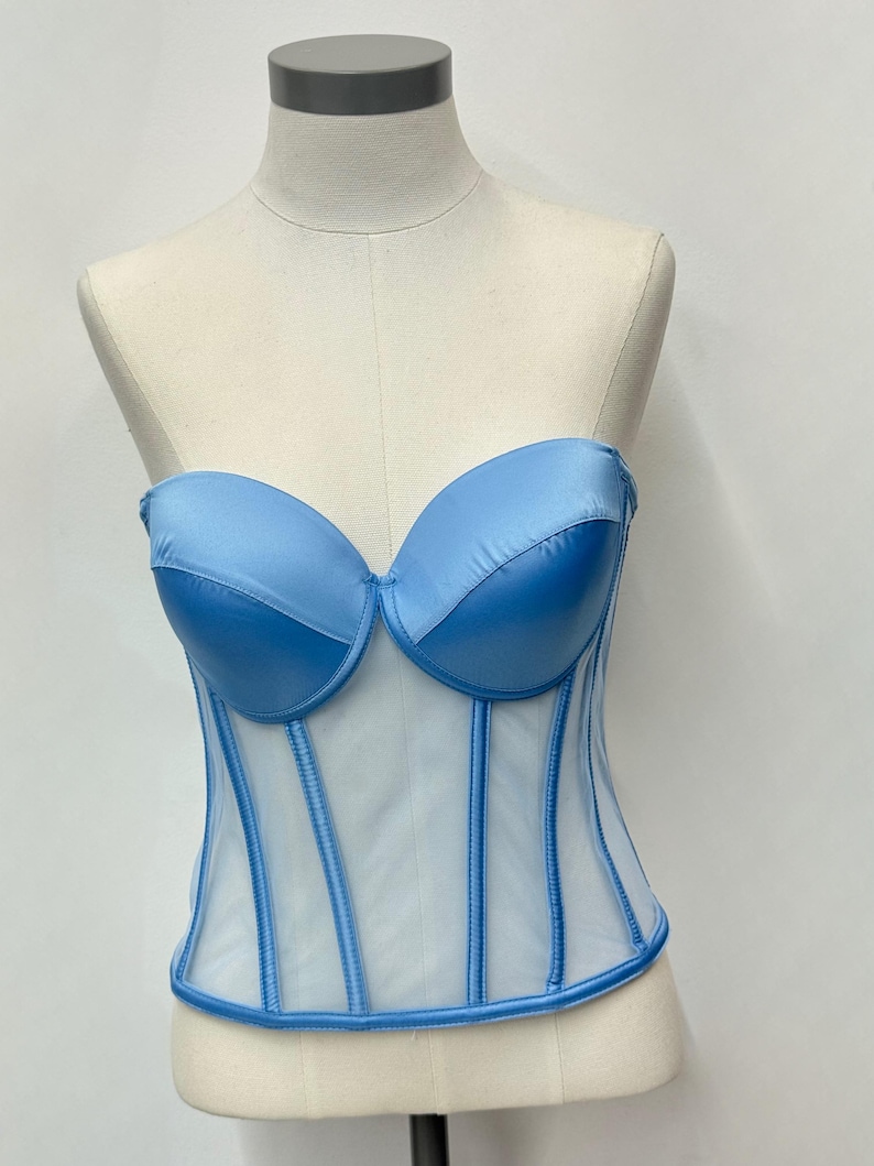 May include: A light blue corset with a sweetheart neckline and boning detail. The bodice is made of a sheer, white fabric with vertical blue accents. The corset is displayed on a mannequin.