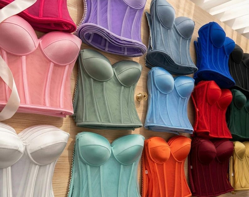 May include: A collection of colorful corsets in various shades, including pink, purple, blue, red, and green. The corsets have a structured design with boning and a sweetheart neckline. Some corsets have a sheer overlay, and others are made of satin.
