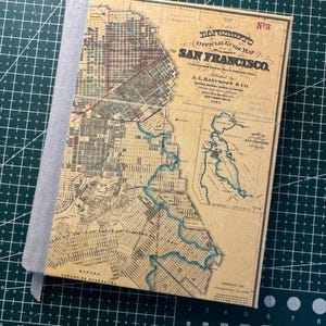 May include: A hardcover notebook featuring a vintage map of San Francisco. The cover displays a detailed map with text that reads "OFFICIAL GUIDE MAP SAN FRANCISCO." The notebook has a gray fabric spine and a ribbon bookmark.