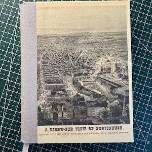 May include: A hardcover book with a vintage black and white aerial view of a city. The book has a light gray spine and a white ribbon bookmark. The title on the cover reads "A Bird's-Eye View of Providence."