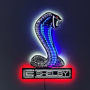 May include: A neon sign depicting a blue cobra above a red and white 'Shelby' logo. The sign is mounted on a dark wall above a television showing a blue sports car. The room has a modern, minimalist design.