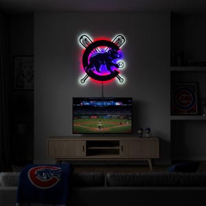 Puede incluir: Un letrero de neón LED montado en la pared con el logotipo de los Chicago Cubs, que consiste en la silueta de un oso azul dentro de una letra C roja, cruzada por dos bates de béisbol. El letrero emite un brillo rojo y azul sobre la pared.