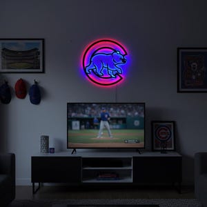 Puede incluir: Un letrero de neón circular que presenta la silueta de un oso azul dentro de una letra C roja. El letrero emite un brillo vibrante contra la pared, mostrando el logotipo de los Chicago Cubs. El diseño está iluminado con tubos de neón azul y rojo.