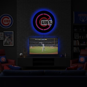 Puede incluir: Un letrero circular de pared con el logotipo de los Chicago Cubs, que presenta una C negra y el texto blanco CUBS, iluminado por un anillo de luz LED azul. El letrero está montado en una pared de ladrillo oscuro sobre una pantalla de televisión.