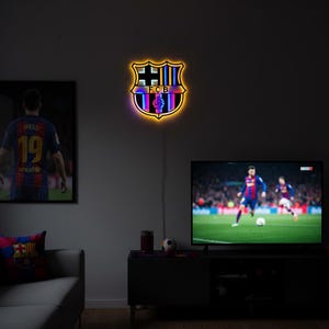 May include: A wall-mounted, shield-shaped FC Barcelona neon sign featuring the club crest with the letters FCB in the center. The sign emits a warm yellow glow around its perimeter, highlighting the blue, red, and yellow colors of the emblem.