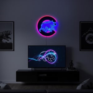 Puede incluir: Un letrero de neón circular montado en la pared con la silueta de un oso azul brillante centrada dentro de una letra C negra, rodeada por un halo de luz rosa vibrante. El letrero está colocado sobre una pantalla de televisión con un oso.
