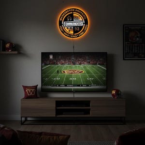 May include: A circular Washington Commanders wall sign featuring a black and gold design with the team logo, the text Washington Football, and the years 1937, 1942, 1983, 1988, 1992, and 32. The sign emits a warm orange glow from behind its circular frame.