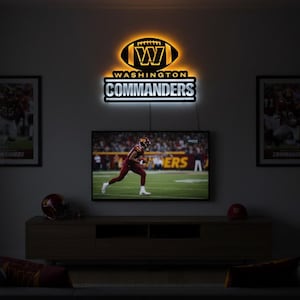 May include: A wall-mounted, illuminated Washington Commanders logo sign featuring a football shape with a central W, positioned above a television screen. The sign emits a warm glow, displaying the team name in bold, white, block lettering.