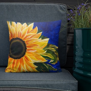 May include: A throw pillow with a vibrant yellow sunflower on a blue background. The sunflower is painted in a realistic style with a dark brown center and yellow petals.